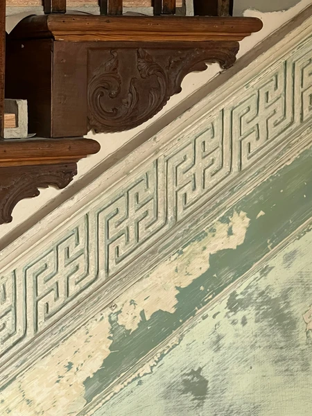 Ornate wooden stair brackets with a Greek key patterned molding on a weathered wall.