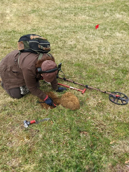 A person kneeling on grass with a metal detector and digging a hole.