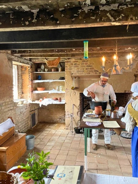 A color photograph of a living history demonstration in a rustic colonial-style kitchen. A man in period-accurate clothing is stirring a large pot over a hearth while another person in a traditional headwrap stands nearby.