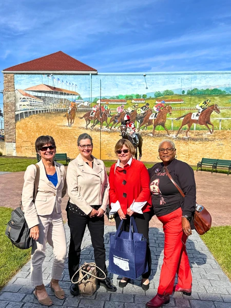 A color photograph of four women standing on a paved path in front of a large outdoor mural depicting a horse race. The mural includes several horses and jockeys in mid-race and a grandstand in the background.
