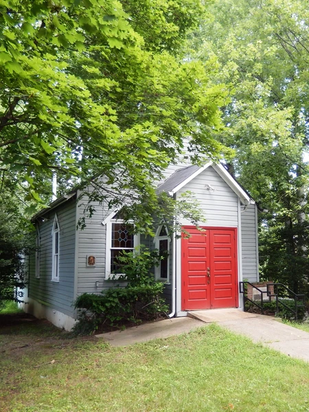 Small gray building with a bright red double door, white trim, and ramp, surrounded by green trees and grass on a sunny day.