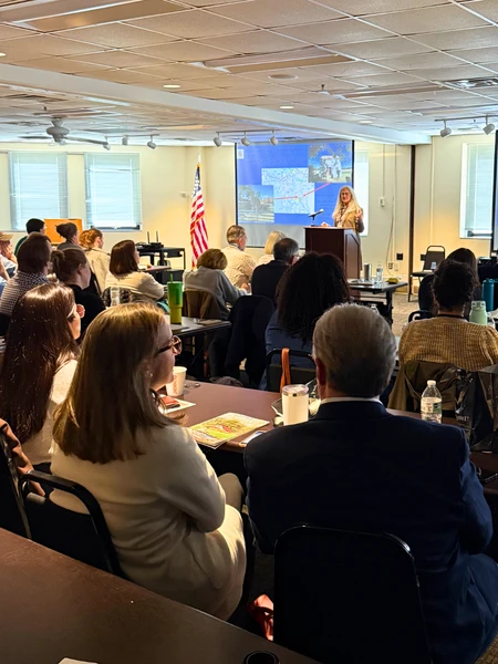 A photo of the audience at a symposium. A woman stands at a podium in front of a projector screen showing a map and historical photos of the Wye Research and Education Center.