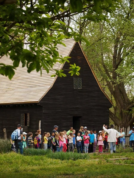 A large group of children listening to a guide in front of a historic dark wood colonial building at the Godiah Spray Tobacco Plantation.