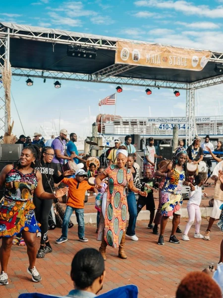 A group of people dancing in colorful attire at the Kunta Kinte Festival in front of an outdoor stage near a harbor.