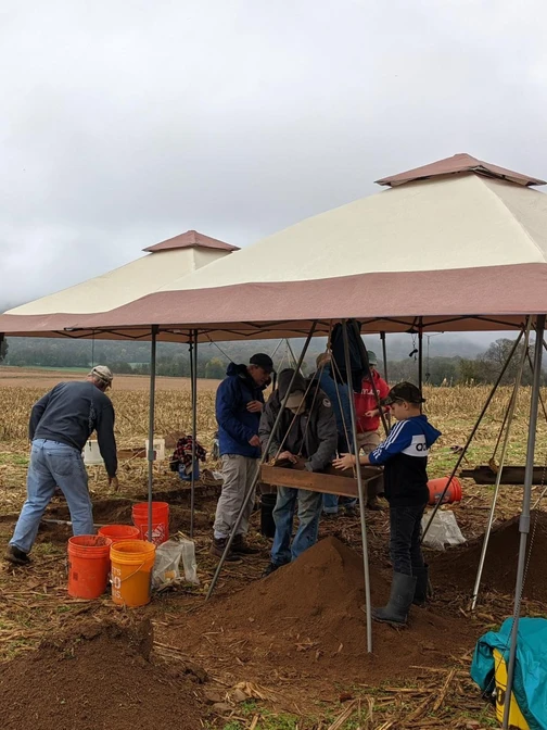 People working at an archaeological dig site in a field under cloudy skies, with equipment and canopies.