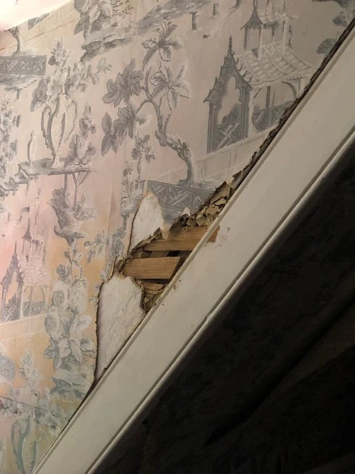 Aged wallpaper with pastoral designs peeling to reveal wooden laths; a window with slatted panes is visible.