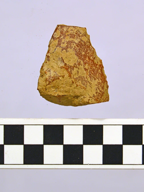 A small, tan and reddish-brown stone flake tool sits above a black and white centimeter scale bar.