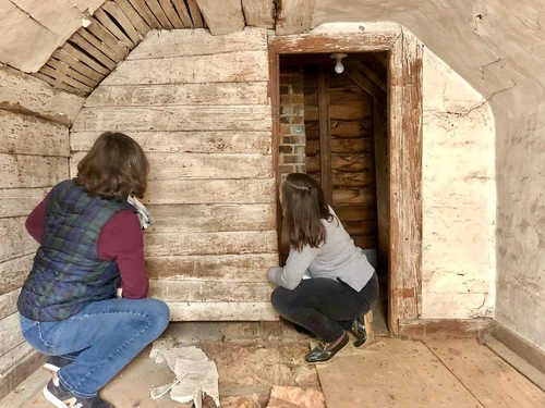 Two preservationists kneel inside a distressed building to inspect a white-washed wooden wall next to a vaulted ceiling with exposed lath.