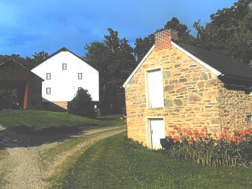 A stone building and barns in a rural landscape with orange flowers and trees under a blue sky.