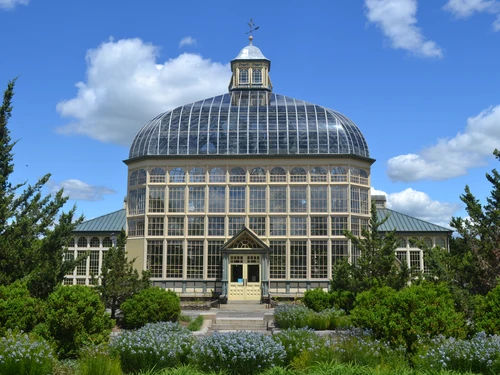 Historic conservatory with a glass dome and numerous windows, surrounded by lush greenery and flowering plants under a blue sky.