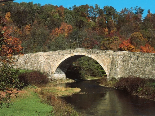 Stone arch bridge spanning a river, framed by trees with fall foliage.