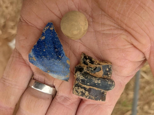 A hand holding a black piece of pottery, a blue fragment, and a small brown ball.