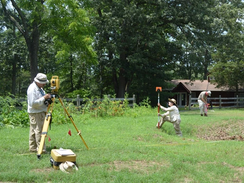 People conducting a land survey in a grassy area surrounded by trees, with survey instruments and small red flags marking the terrain.
