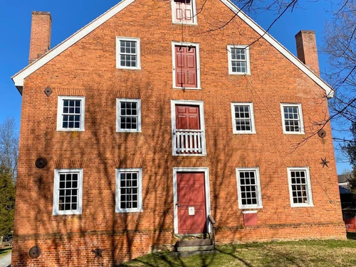 A three-story brick building with red doors and white-trimmed windows, casting tree shadows on its facade under a clear blue sky.