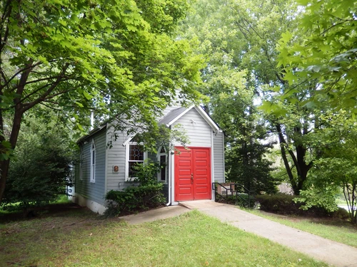 Small gray building with a bright red double door, white trim, and ramp, surrounded by green trees and grass on a sunny day.