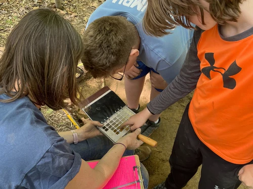 An adult and two children examine a soil color chart at an outdoor excavation site.