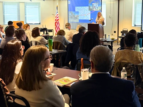 A photo of the audience at a symposium. A woman stands at a podium in front of a projector screen showing a map and historical photos of the Wye Research and Education Center.