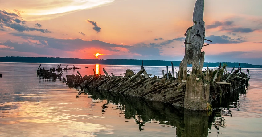 A vibrant sunset photograph over Mallows Bay in Maryland, featuring the skeletal wooden remains of a sunken ship rising from the calm water.