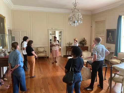 A group of people standing in an elegant room with wooden floors and a chandelier, listening to a speaker.