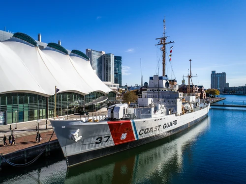 Historic Coast Guard ship, docked near a modern building with a wavy white roof. City skyline and calm water reflect a clear blue sky.