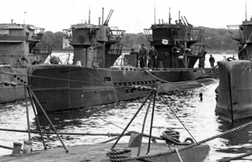 Several German U-boats docked in a harbor with the central sub features a large conning tower with crew members visible on deck.