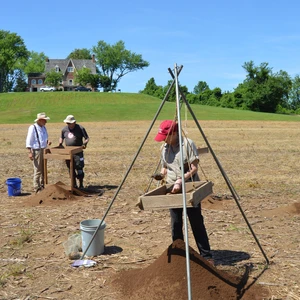 Three people conduct an archaeological dig in a field, using screens to sift soil. A historic house and lush trees are visible on a distant hill.