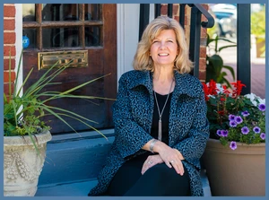 A color portrait of a woman smiling while sitting on the blue-painted steps of a historic brick building, flanked by colorful potted flowers.