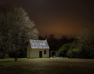 A solitary white cottage at night under a cloudy orange-lit sky, surrounded by bare trees and lush greenery, evokes a quiet, eerie atmosphere.