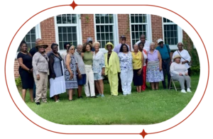 A large, smiling group of Black adults and elders pose together on a lawn in front of a brick building with white windows.