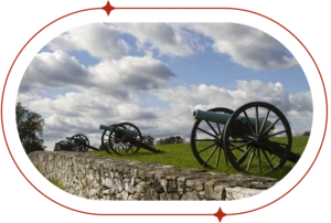 Row of Civil War-era cannons sitting on green grass against a blue and cloudy sky.