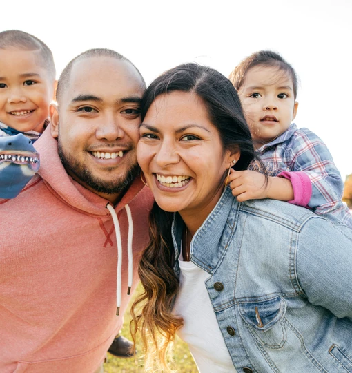 A medium, outdoor shot of a family of four smiling for the camera. The two young children are carried piggyback