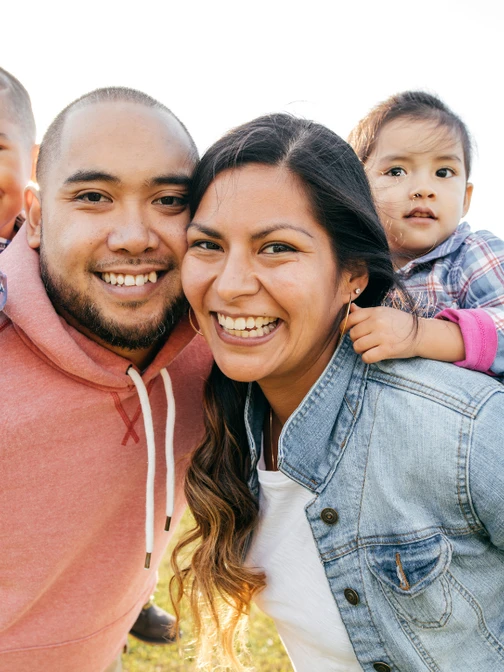 A medium, outdoor shot of a family of four smiling for the camera. The two young children are carried piggyback