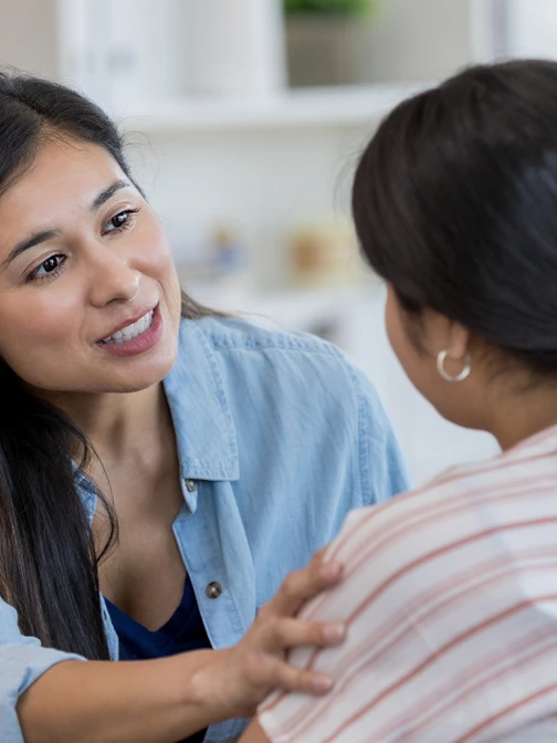 A medium indoor shot of two women on a couch. One woman with dark hair, wearing a denim shirt, is speaking to the other woman, who has her back to the camera, while gently touching her shoulder.