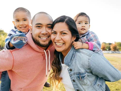 A medium, outdoor shot of a family of four smiling for the camera. The two young children are carried piggyback