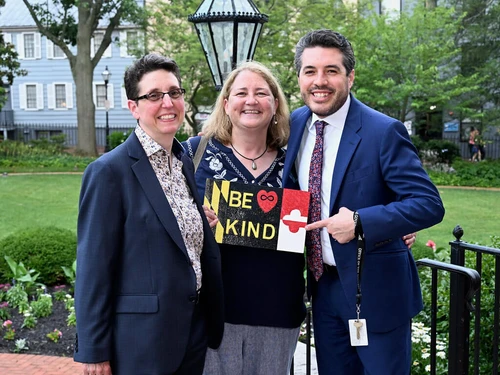Three people standing together with one pointing to a sign that reads "Be Kind"