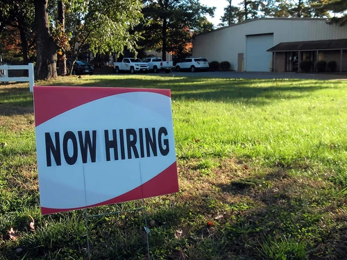 A "Now Hiring" sign on a green front lawn
